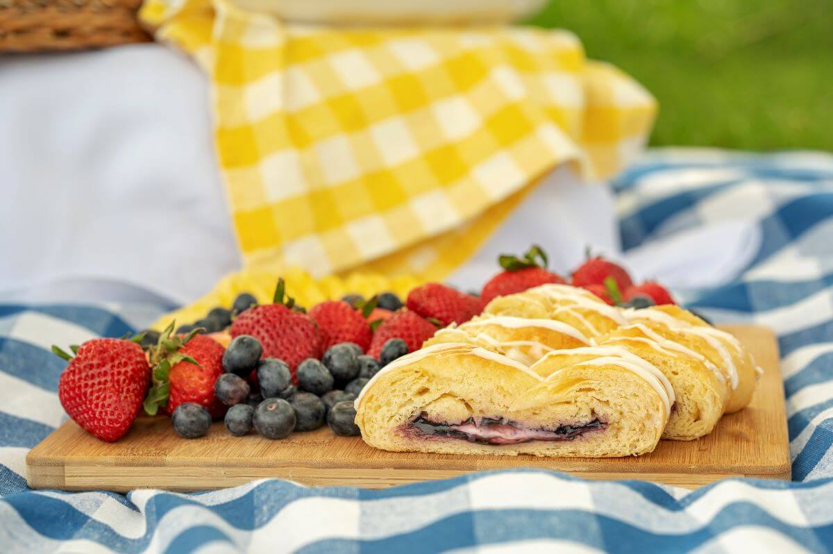 Slices of a Blueberry & Cream Cheese pastry on a wood cutting board along with several fruits. The board is on a picnic blanket.